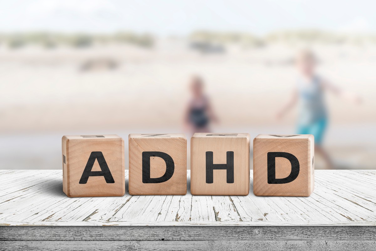 Wooden letter blocks spelling out the letters ADHD with a sandy beach scene in the background where two children are playing