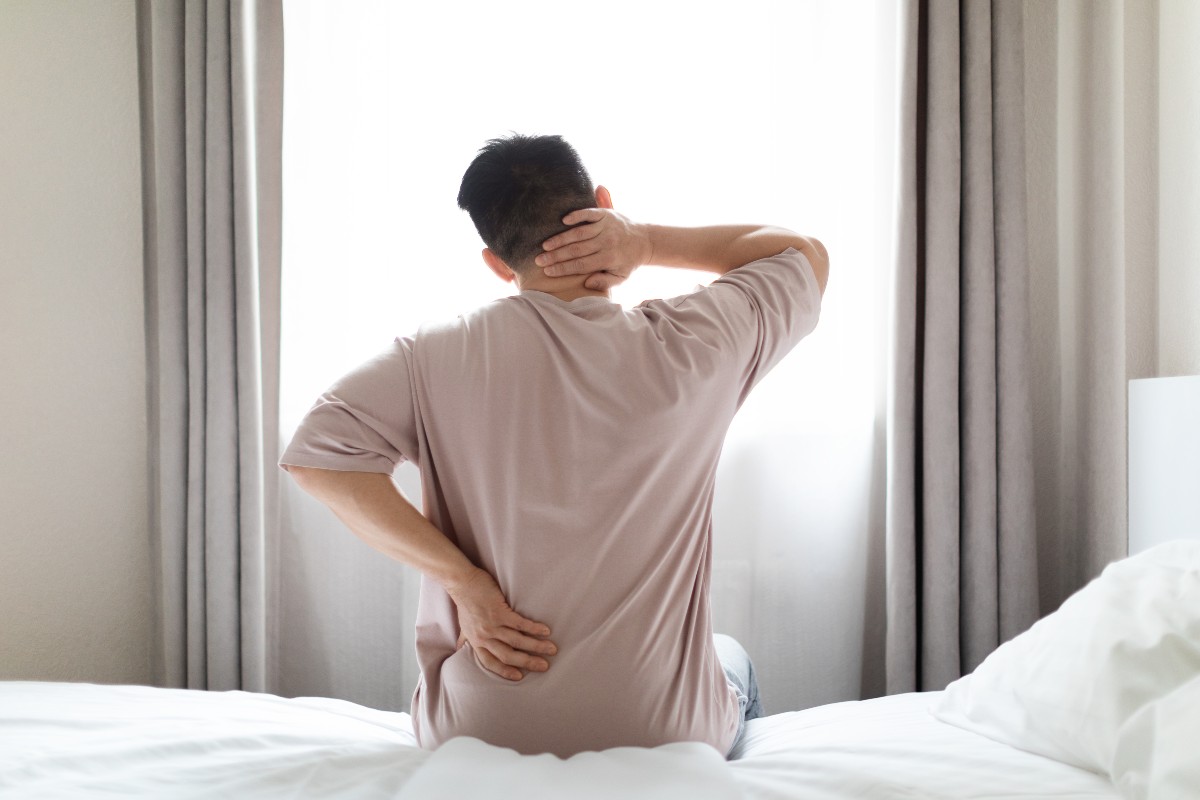 A man sat on the edge of his bed looking out of his bedroom window while rubbing his back and neck to soothe them