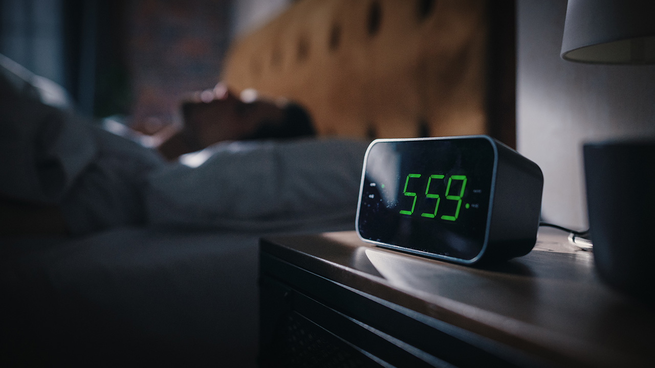 A man asleep in bed with an alarm clock that reads 5:59am in the forefront of the image