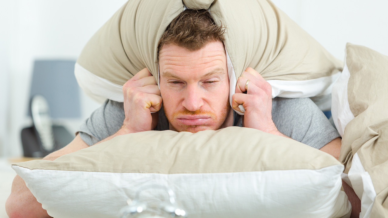 A man looking grumpy and fed up as he wakes up in the morning. He is covering his ears with his pillow to try and block out the noise.