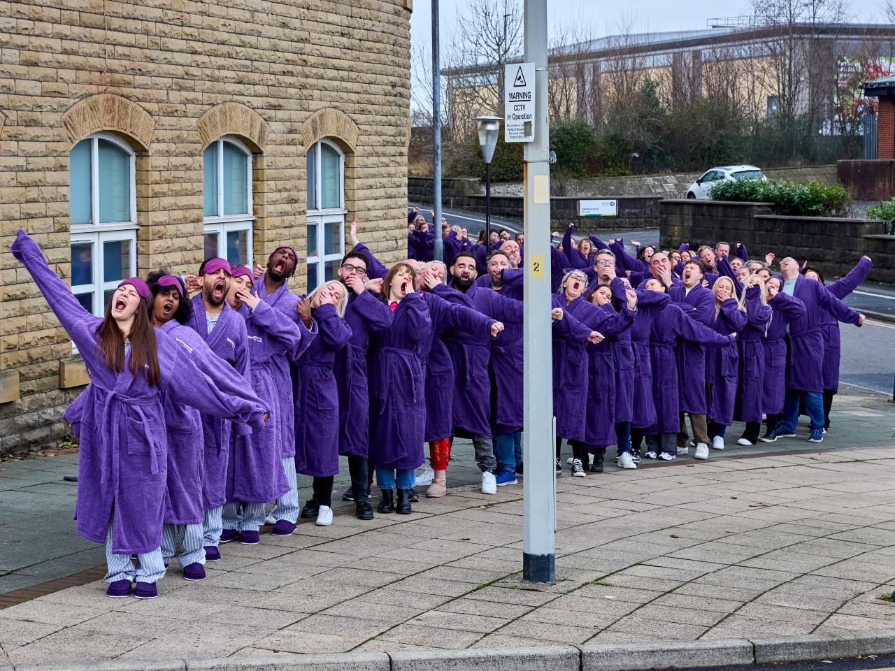 A long line of yawning Bensons for Beds staff members outside of the Bensons for Beds Head Office wearing purple dressing gowns.