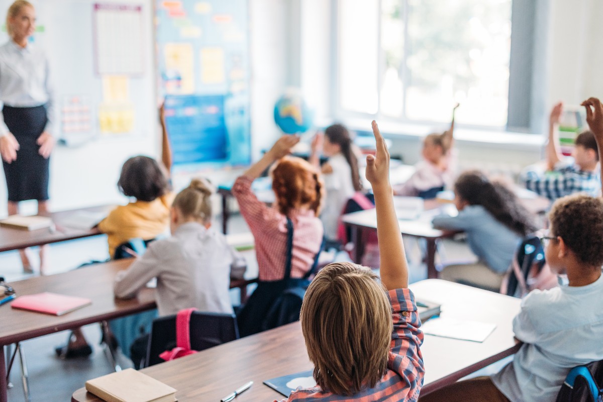 A classroom in full swing, with children sat at desks, several with arms raised and the teacher smiling enthusiastically at the front of the classroom.