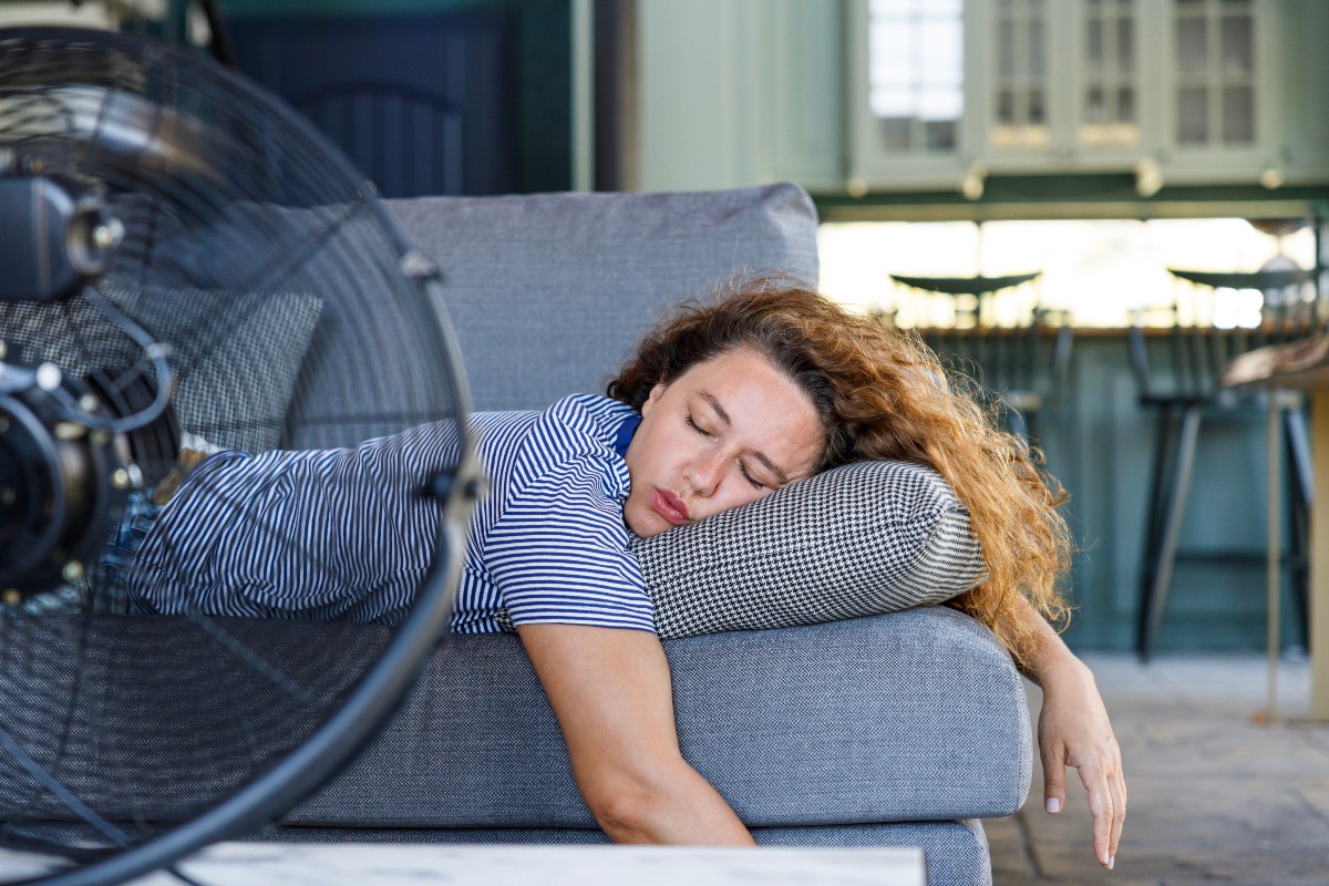 A woman asleep on her front on a couch, a breakfast bar behind her and a fan positioned on top of a coffee table in front of her