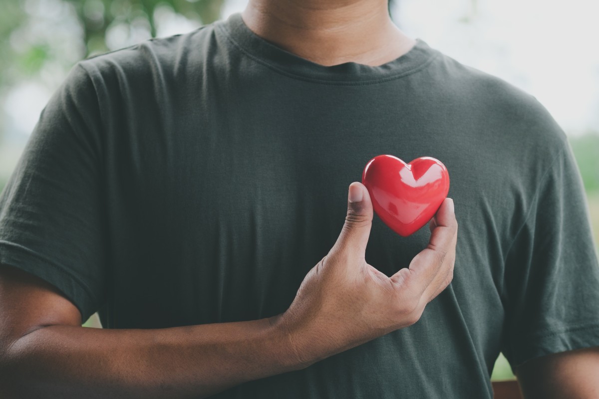 A close up of a man's chest, his right arm holding a red heart just to the left of his chest.