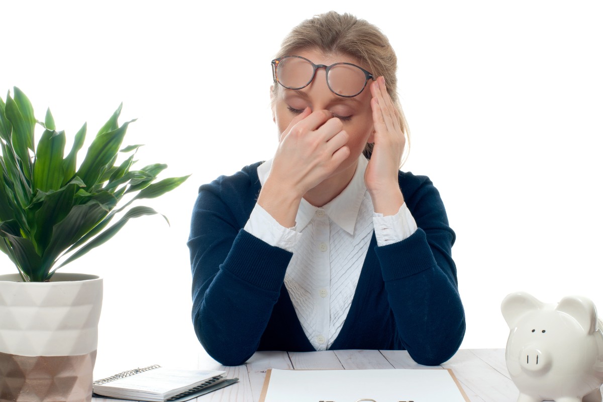 A woman sat at a desk with a plant on one side of her and a piggy bank on the other, she is rubbing the bridge of her nose with her eyes closed
