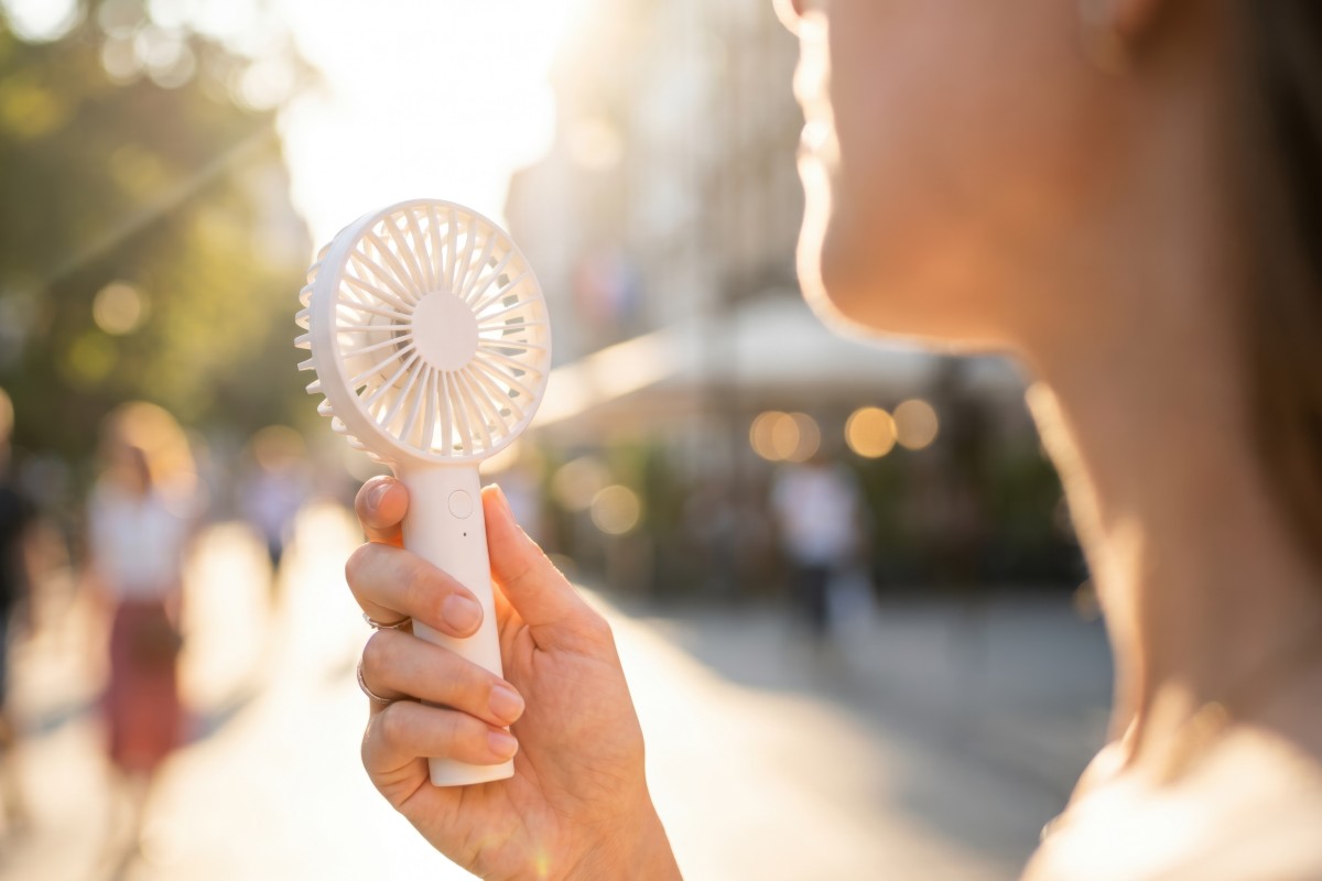 A close up of a woman with a handheld personal fan held up to her face, she is outdoors, walking in the sun during warm weather