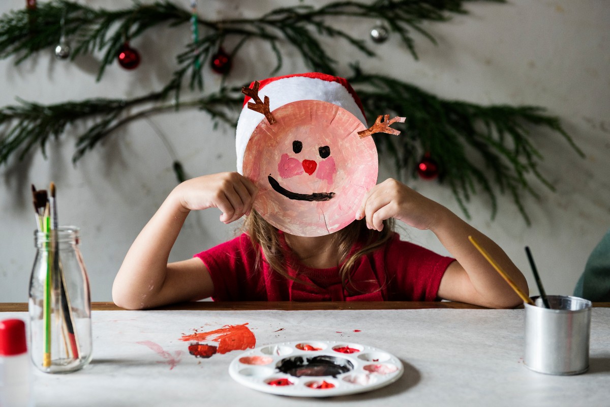 A child holding a decorated paper plate face up in front of their face while sat at a crafting table with a Christmas tree in the background.