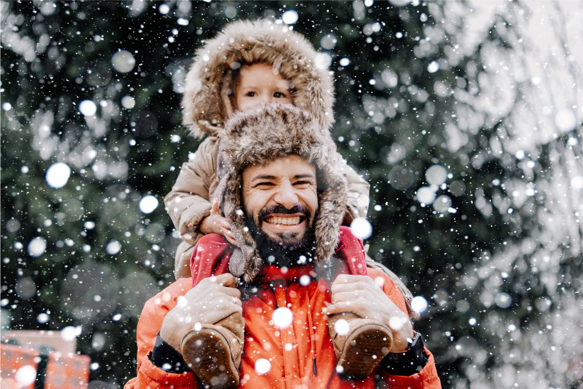 A man with a child on his shoulders walking through the snow with a happy smile on his face