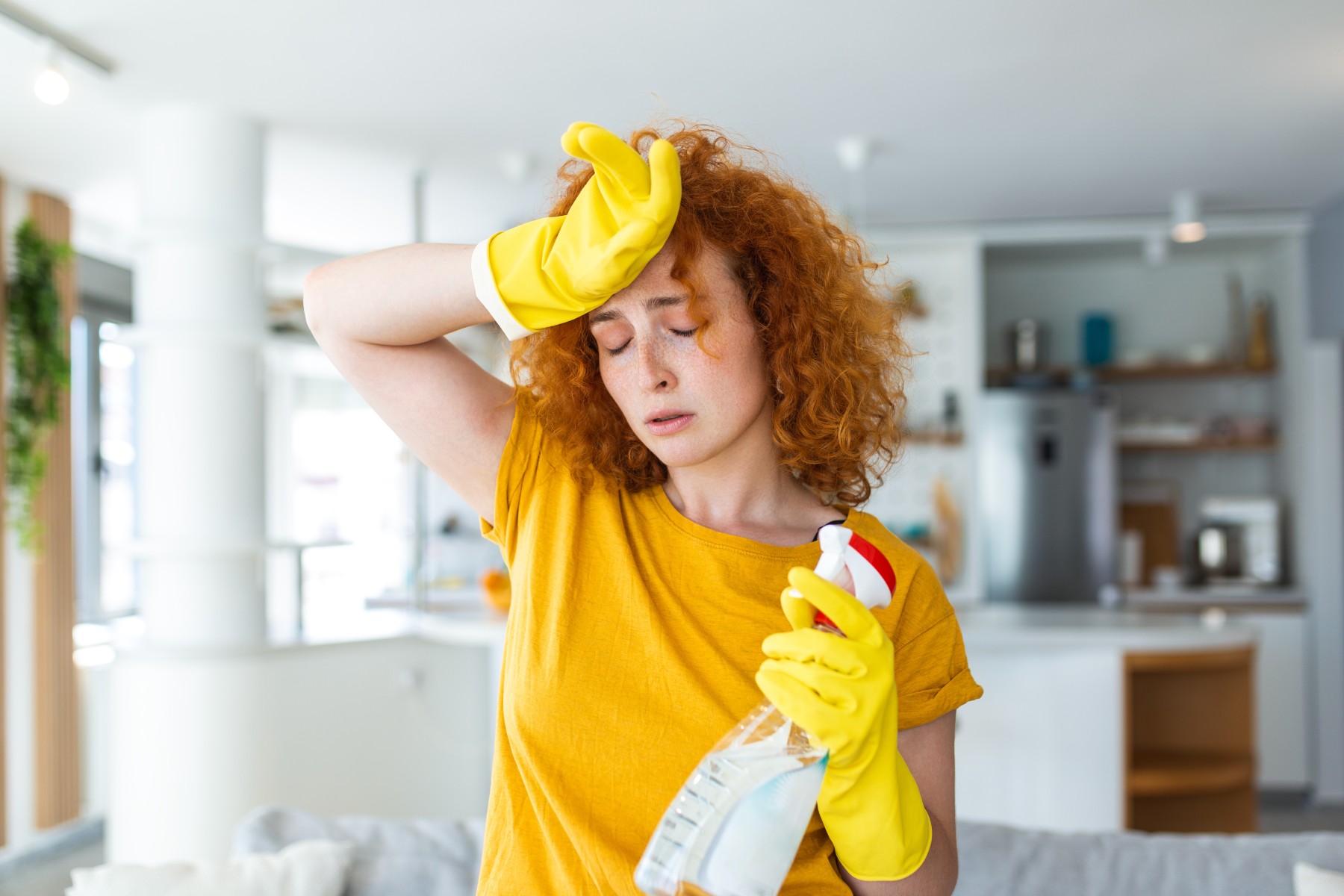 A woman wiping her brow while wearing yellow rubber gloves and holding a bottle of cleaning spray