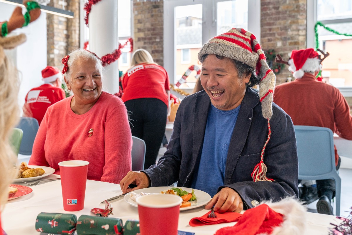 A man and a woman sat enjoying a Christmas meal, smiling, wearing Christmas hats, and having a good time.