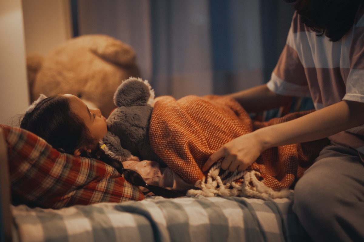 A warm bedtime scene with a child cuddling a soft toy while an adult tucks them in.
