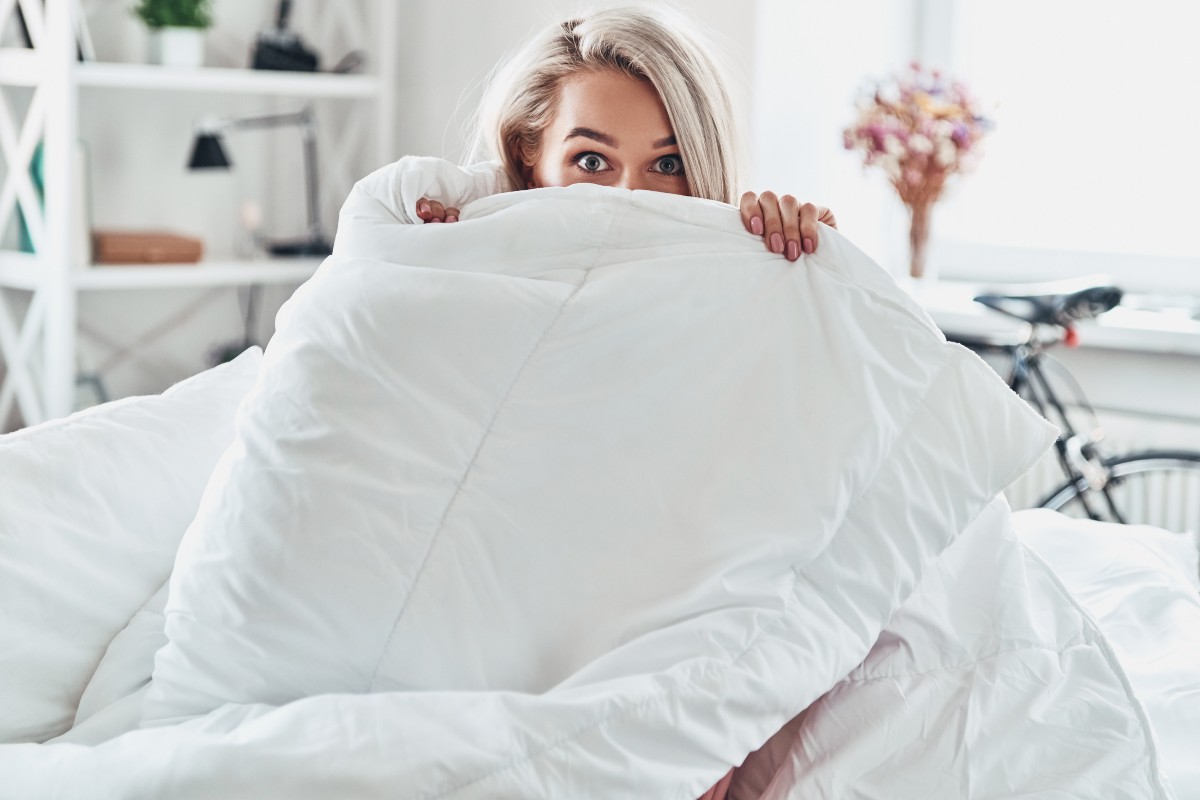 A woman sitting on a bed, mostly wrapped in her duvet with just her eyes, fingers, and the top of her head peeking over the top of the duvet.