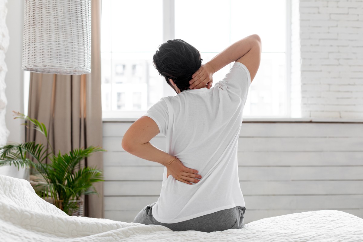 A person sat on a bed with their back to the camera, rubbing their back and neck after sleeping on a mattress that is too soft.