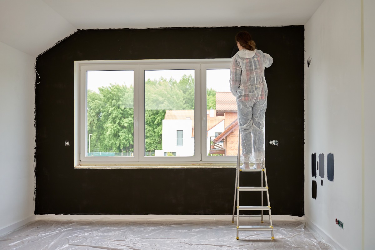 A lady painting a feature wall on her picture window bedroom wall, balanced on a step ladder