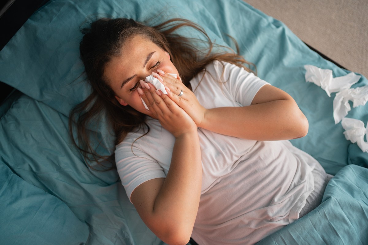 A woman lying on her back in bed with a tissue to her nose as she blows it; beside her is a pile of crumpled used tissues