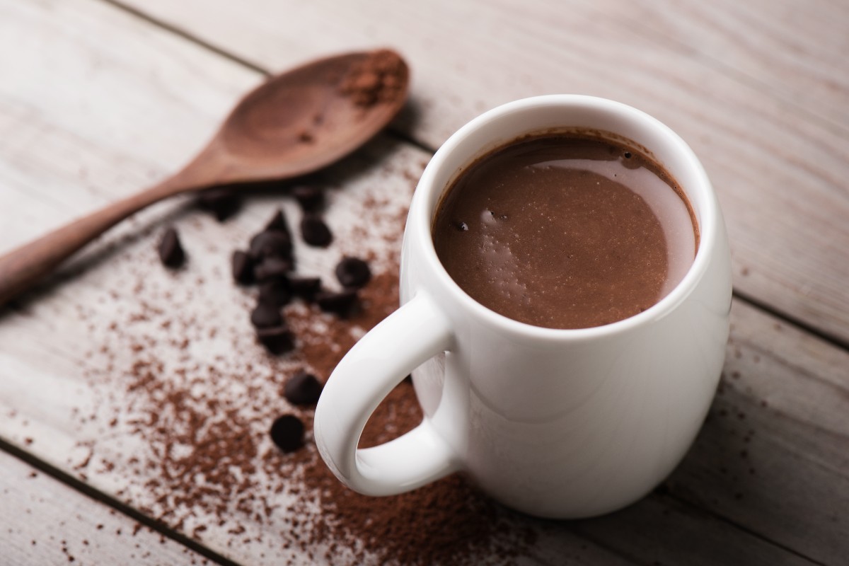 A mug of hot chocolate on a wooden table top beside some cocoa powder and a wooden spoon.