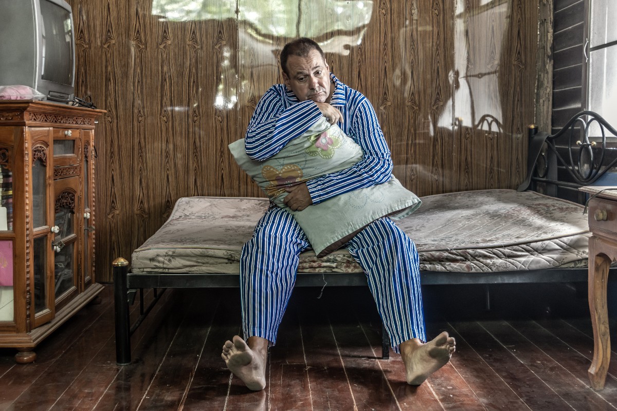 A lonely man sat on an old and dirty mattress in his bedroom, clutching his pillow to his chest with a sad look on his face.