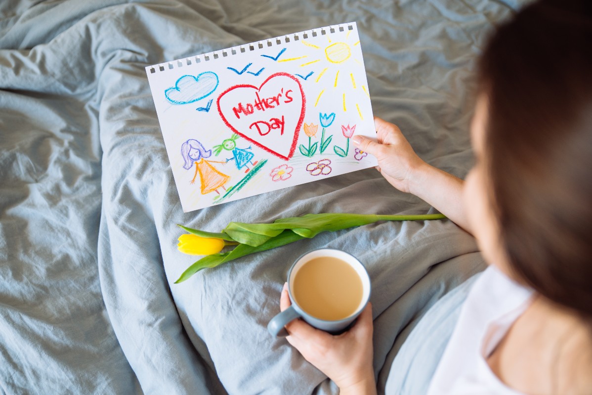 A mum sat in bed with a cup of tea in one hand and a picture drawn by her child in the other.