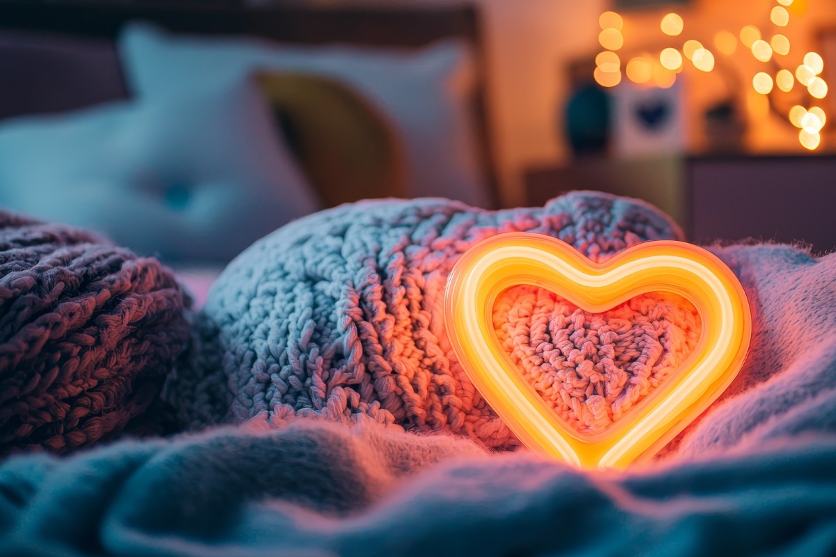 A heart nightlight glowing in the dark on a child's bed. There are also warm coloured fairy lights glowing in the background on the bedside table.