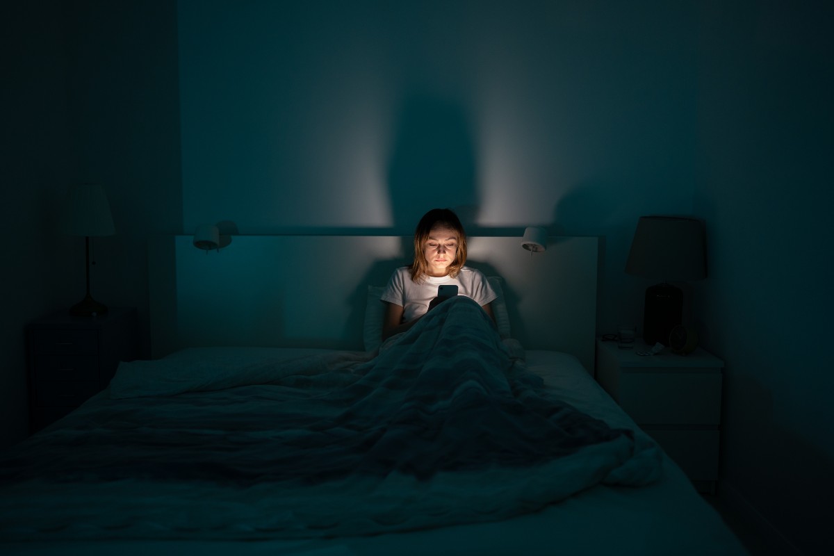 A lady sat up in bed looking at her phone in an otherwise dark room.