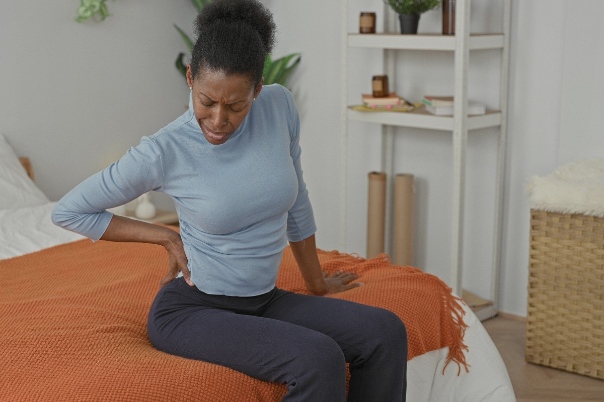 Someone sitting on a bed and holding their lower back in discomfort.