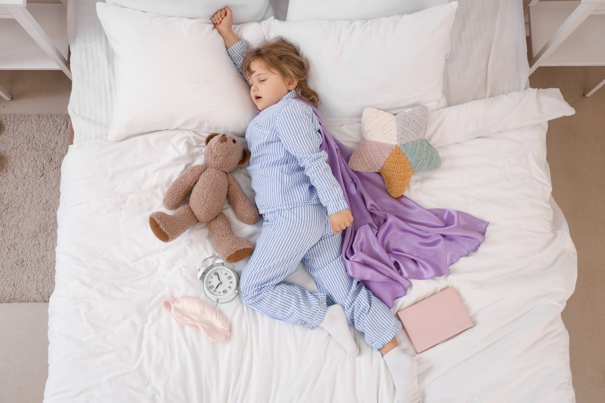 A child asleep in bed with one arm raised above their head in a superhero pose, a cape made from a blanket trails behind them and a teddy is nestled close on the other side of them.