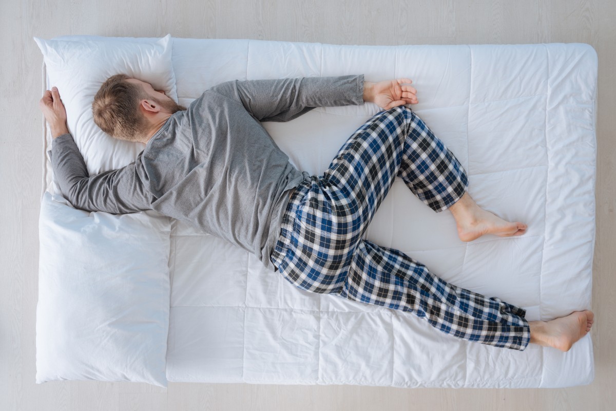 A man asleep on his front, face planting the pillow with one arm raised above his head, his head and neck twisted to one side.