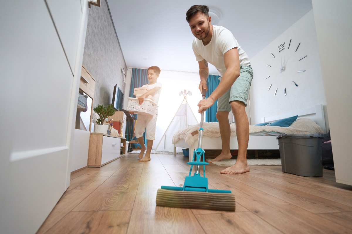 A dad and son spring cleaning their bedroom together: the young boy is organising his toys while dad mops the floor.