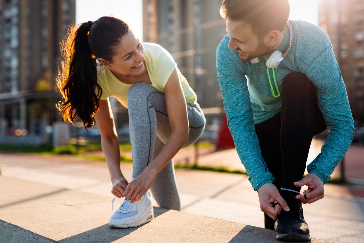 A man and woman lacing up their running shoes outdoors looking at each other and smiling.