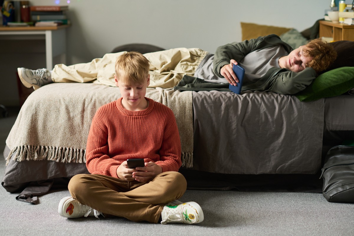 2 brothers playing on screens, one sat crossed legged on the floor, the other lying on the bed.