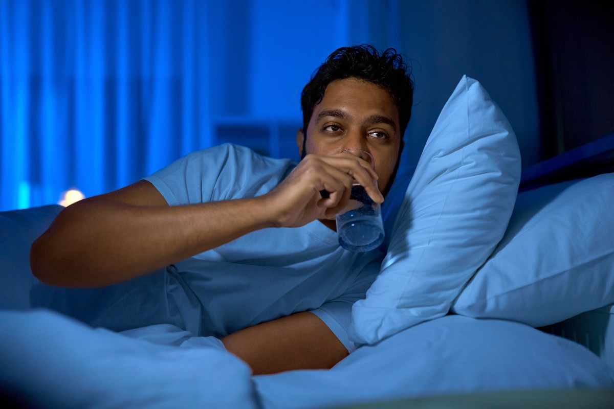 A man drinking from a glass of water in bed after waking up due to dehydration.