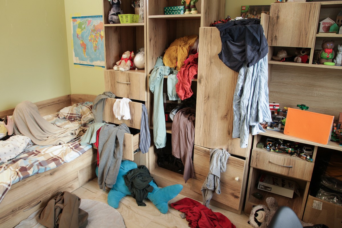 A chaotic wardrobe with clothes spilling out of the wardrobe doors and drawers onto the floor.