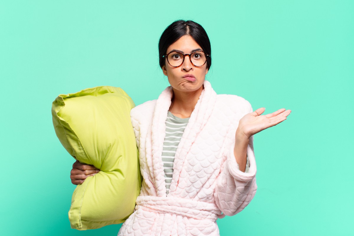 A woman wearing a dressing gown clutching a pillow with one arm and pulling a shrug with the other, her face reads confusion over what to wear to bed.