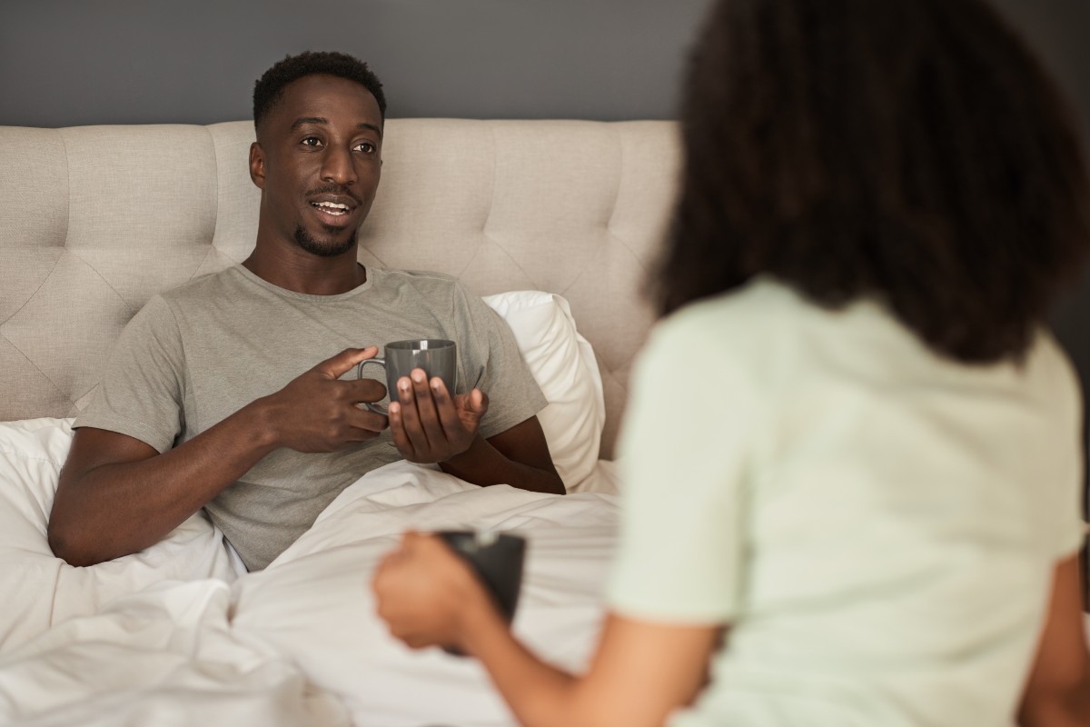 A couple chatting in bed, both holding a mug.