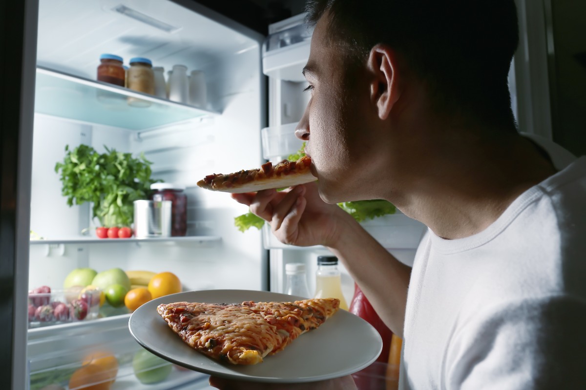 A man eating a slice of pizza at night, illuminated only by the open fridge in front of him.