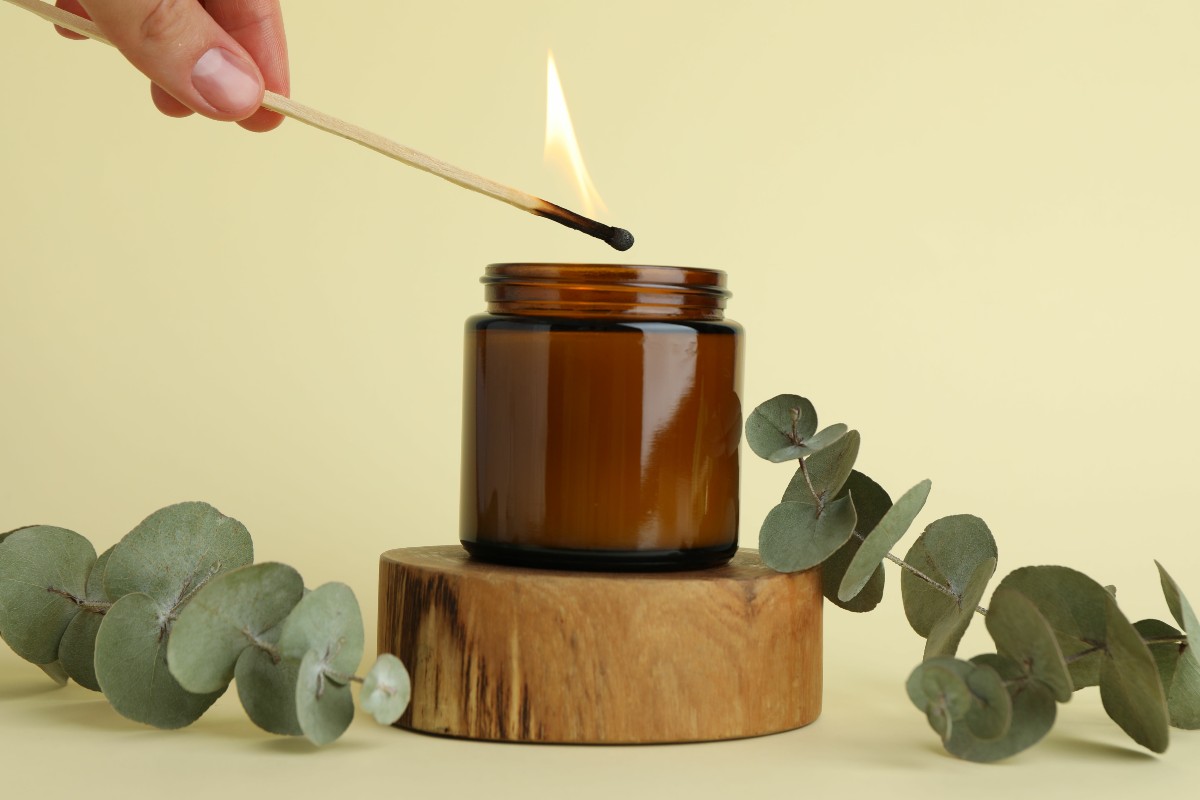 A hand lighting a candle in a deep brown glass jar with some green eucalyptus leaves on either side all set against a soft yellow painted wall.